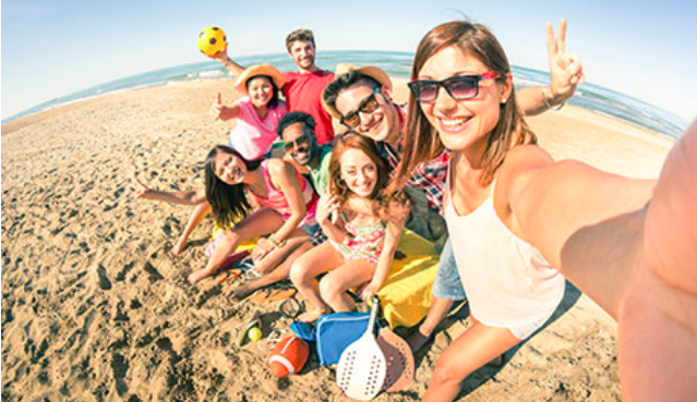 Seven travelers smile in beach attire while taking a beach selfie. 