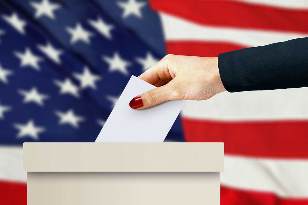 person dropping a ballot in a box in front of an American flag.