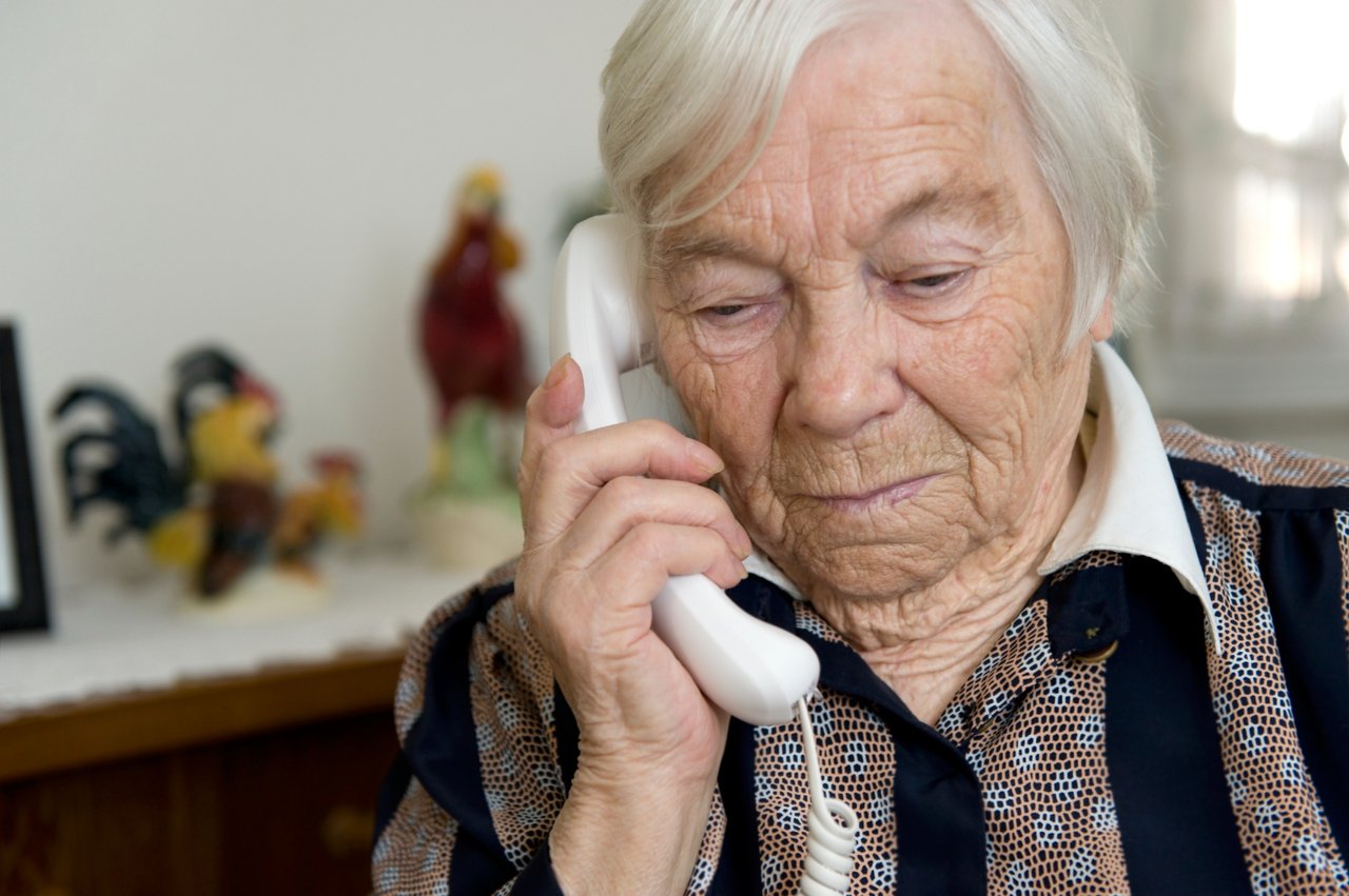 A senior lady holds a corded phone up to her ear in her living room.
